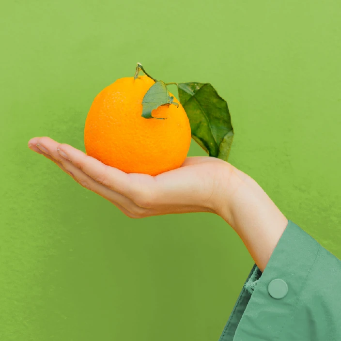 A hand holding an orange in front of a bright green wall
